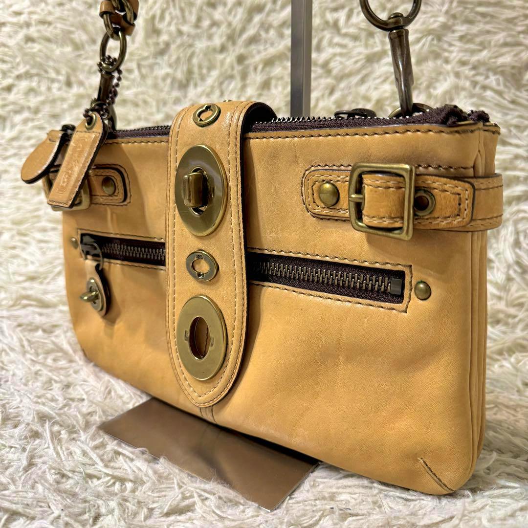 Tan Coach Legacy one-shoulder cow leather bag with turnlock hardware and a front zipper pocket shown on a stand.