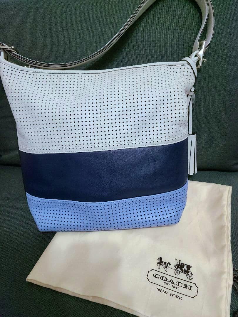 Front view of the COACH Legacy perforated rugby stripe leather duffle bag in white, navy, and light blue placed on a COACH dust bag.