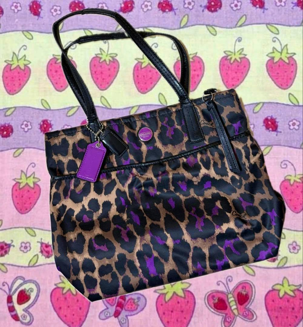 COACH leopard-print canvas shoulder bag with black straps and purple accents photographed on a patterned fabric background.