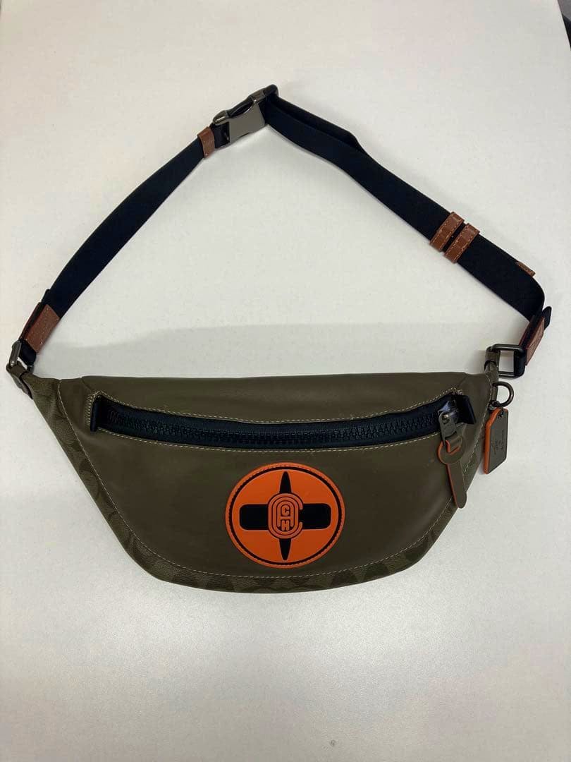 Front view of the canvas body bag described, showing an olive-green bag with an orange circular patch, a black zipper, and an adjustable black strap on a white background.