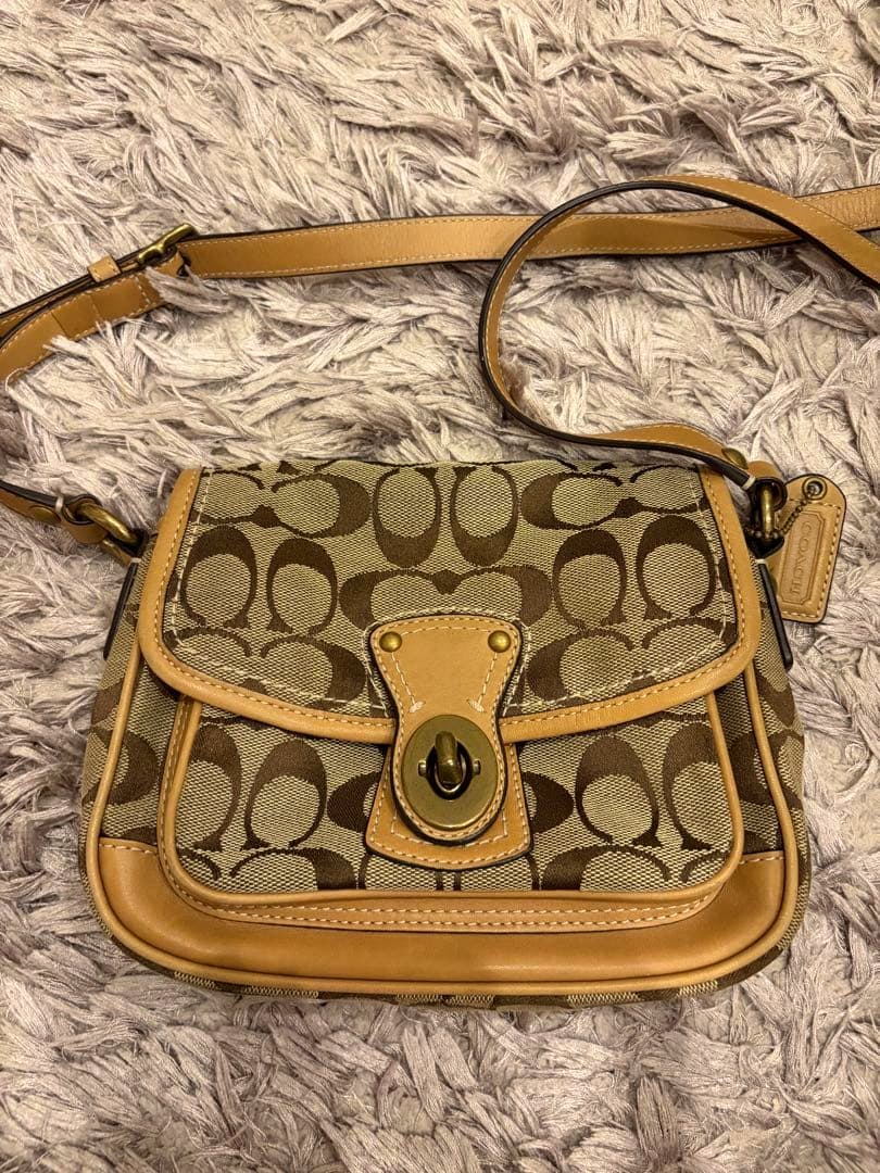 Front view of a Coach logo-pattern canvas shoulder bag with tan leather trim and a turn-lock closure placed on a carpet.