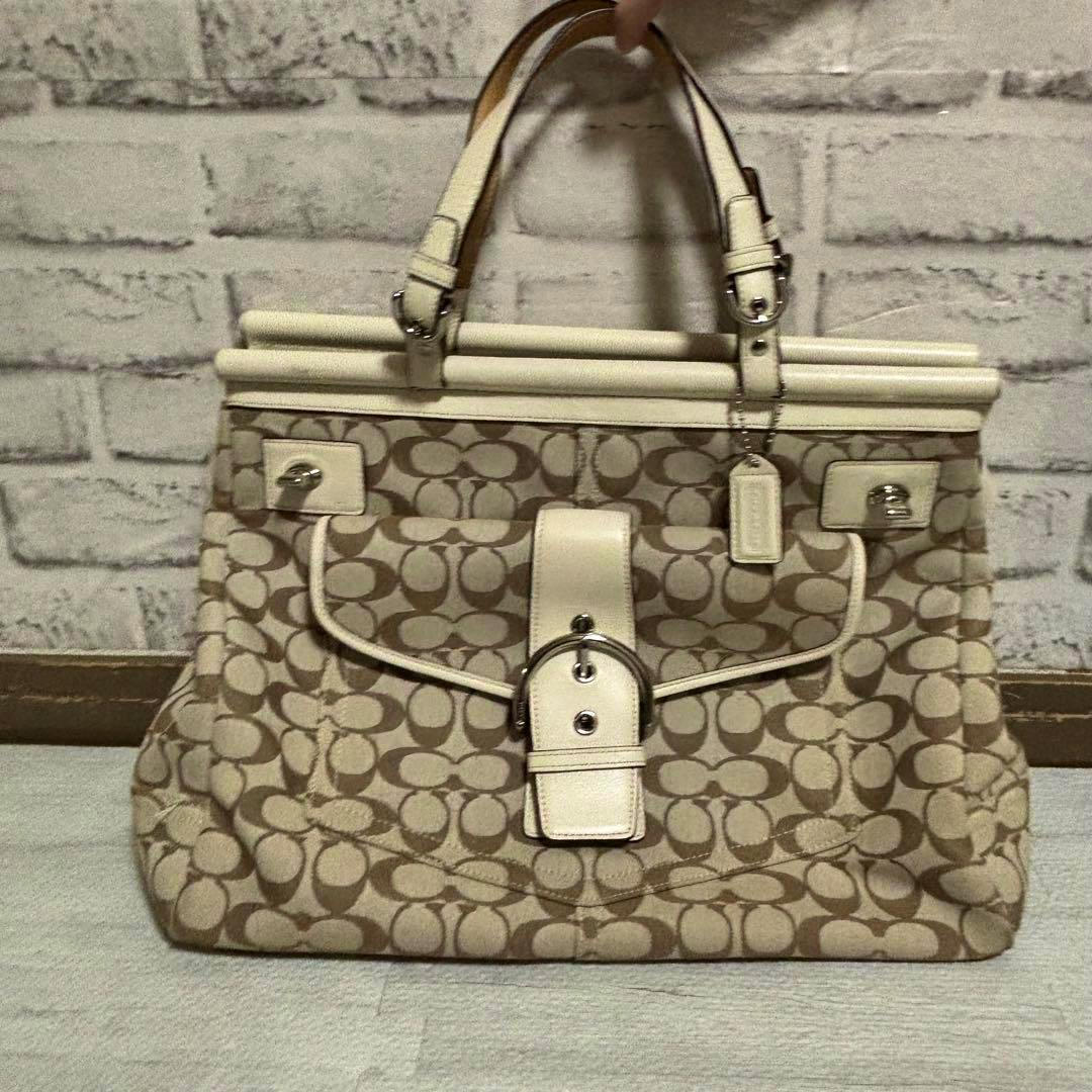 Front view of a beige and white Coach logo-pattern canvas handbag with a buckle strap and top handles.