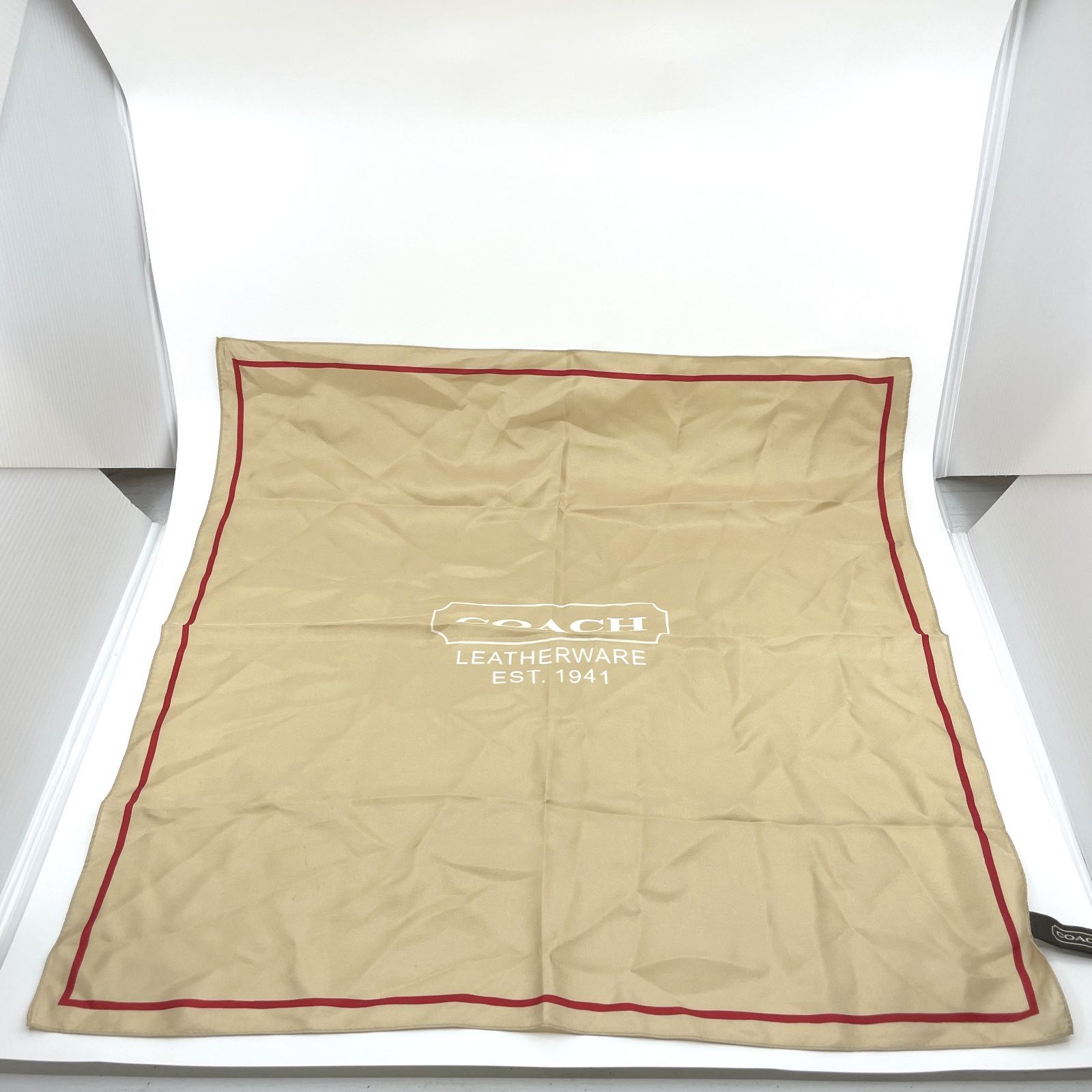 Beige Coach silk scarf laid flat showing the center Coach logo and a red border.