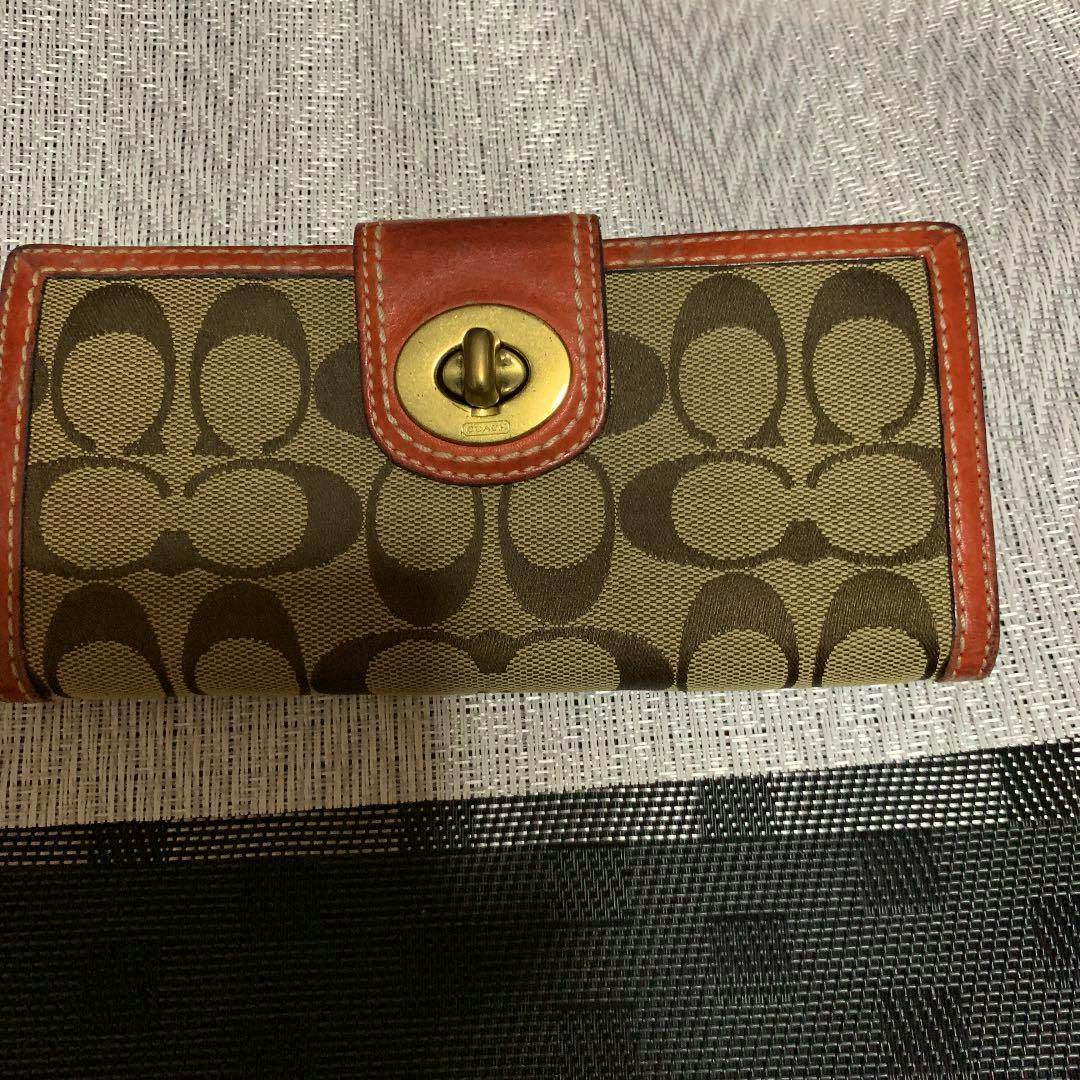 A COACH long canvas wallet showing the brown 'C' monogram pattern and tan leather trim on a textured surface.