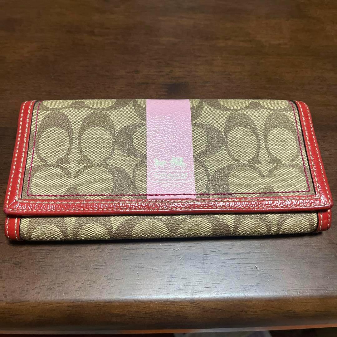 Folded COACH long canvas wallet with signature C pattern, a pink center stripe, and red trim photographed on a wooden surface.