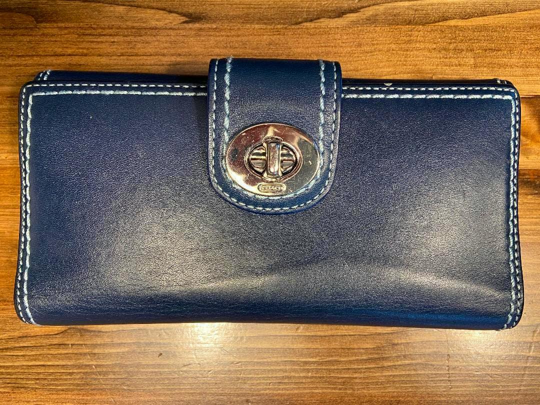 Closed navy leather long wallet showing the front with a turn-lock clasp and contrast white stitching on a wooden surface.