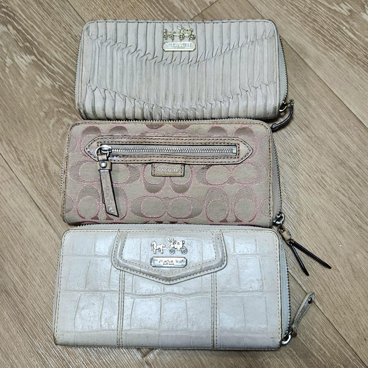 Three Coach long zip-around wallets with different exterior textures and logos placed on a wooden floor.