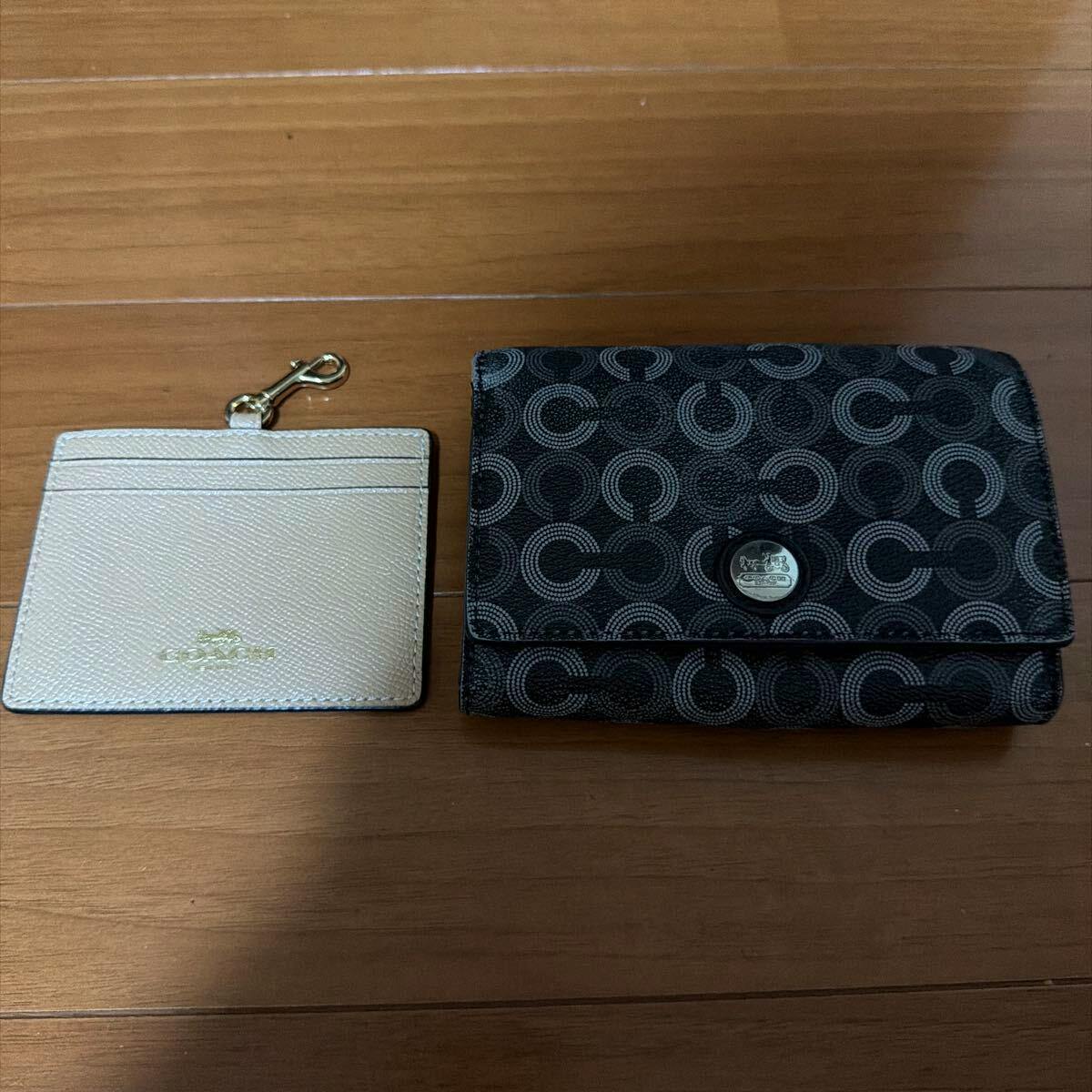 A black patterned Coach wallet and a light-colored Coach pass case placed side by side on a wooden floor.