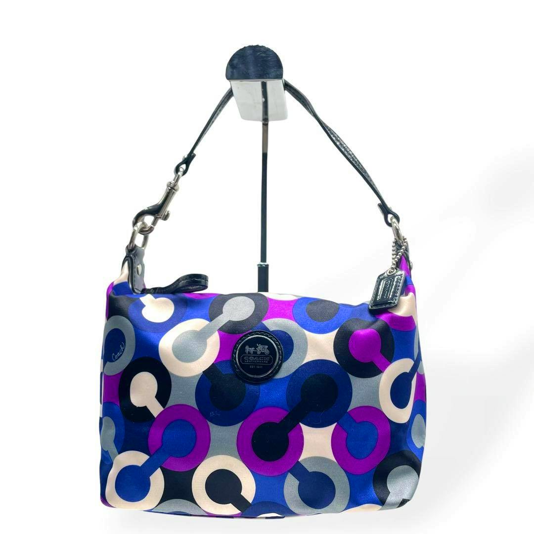 Front view of a COACH Madison graphic op-art accessory pouch with blue, purple, gray and cream circular pattern, black leather strap, and round Coach medallion.