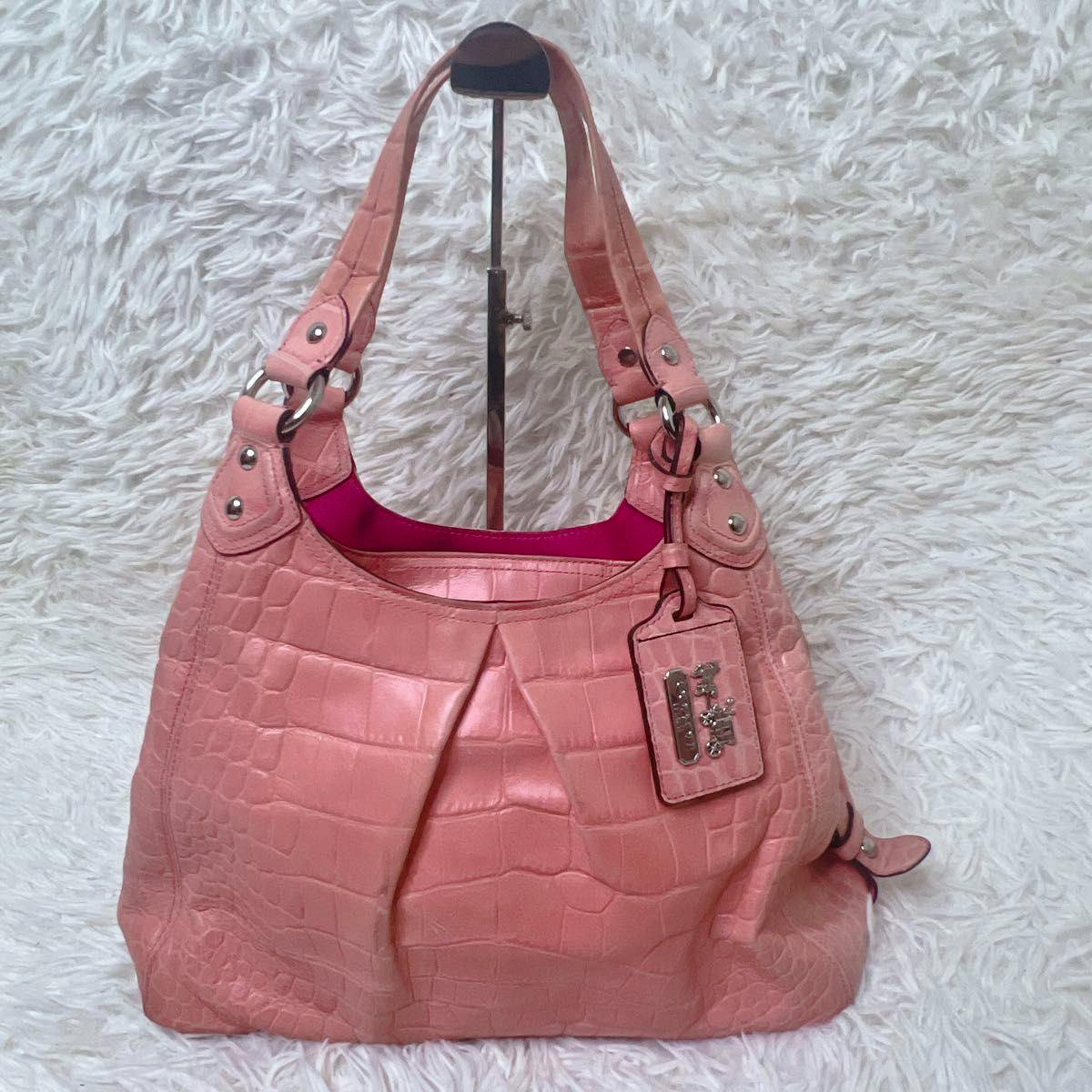 Front view of the pink croc-embossed leather COACH shoulder bag with dual straps and an attached hangtag on a white shag background.