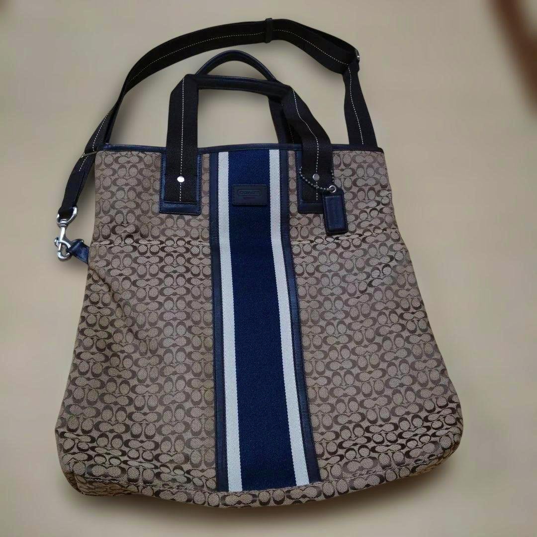 COACH men's 2-way canvas bag with brown monogram pattern and a central navy-and-white vertical stripe, shown flat with handles and shoulder strap.