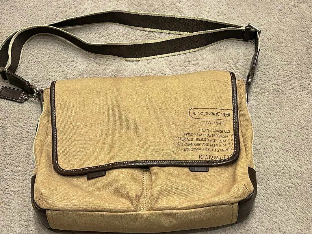 Beige COACH men's canvas shoulder bag with a front flap, brown trim, and an adjustable shoulder strap laid flat on a soft surface.