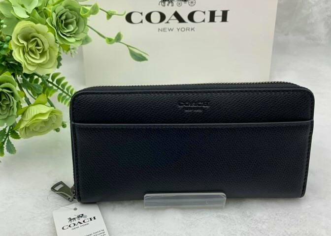 Front view of a black COACH long zip men's wallet (canvas) displayed with its tag and a Coach box behind it.