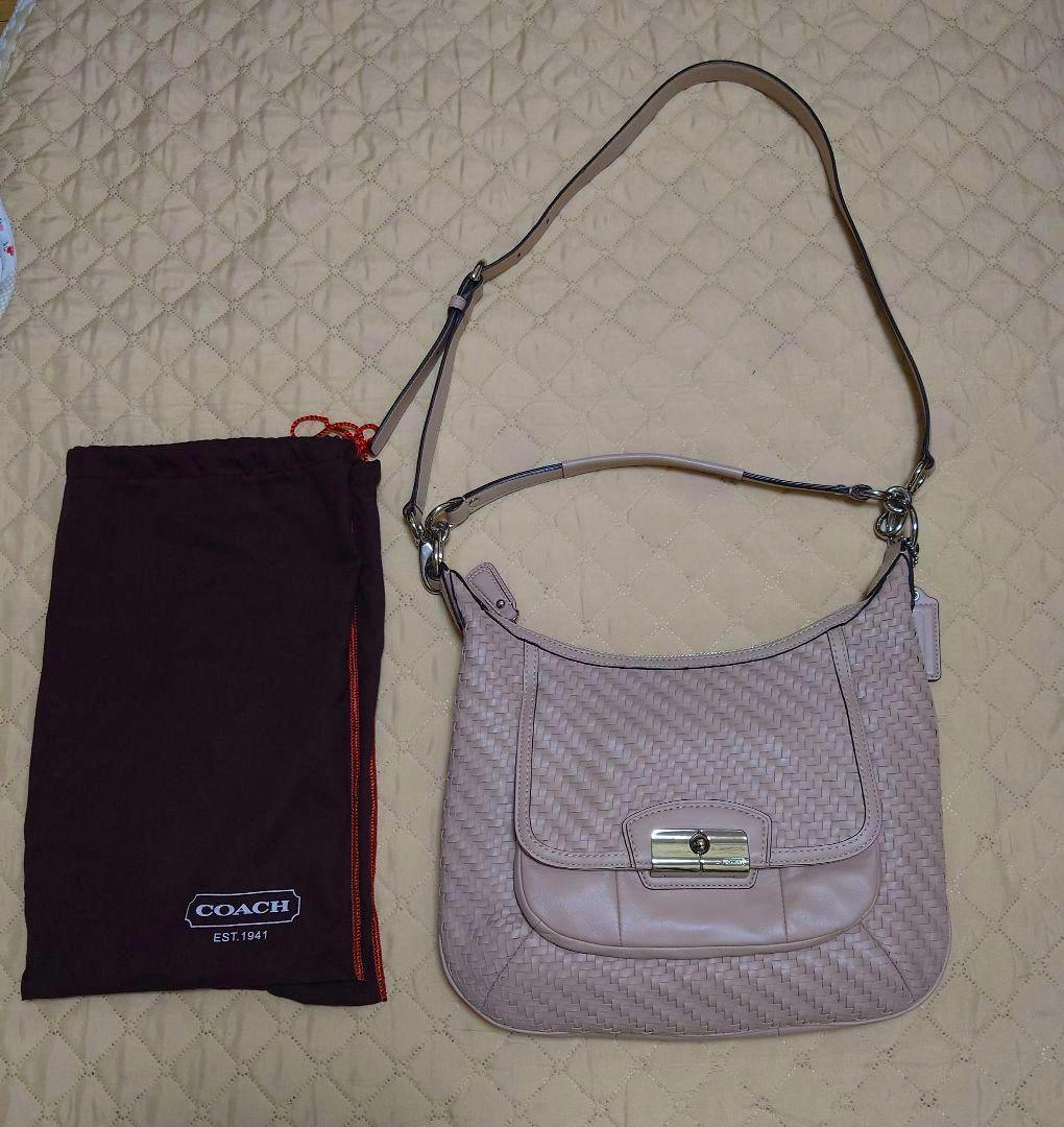 Light pink woven Coach 2-way shoulder bag with a long strap placed next to a brown Coach dust bag on a quilted surface.