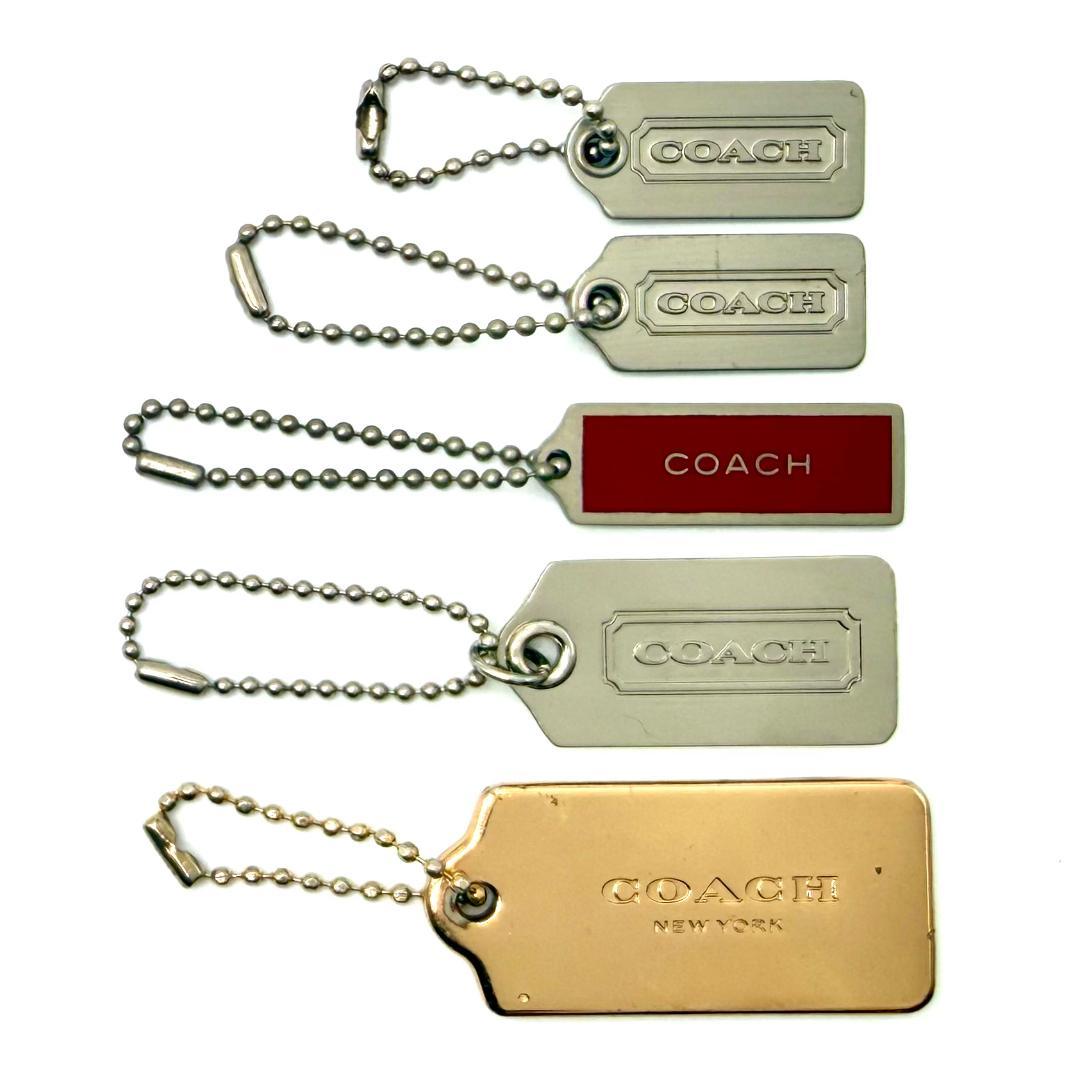 Five Coach metal bag charms in silver and gold tones arranged vertically, each with a ball-chain attachment and Coach branding.