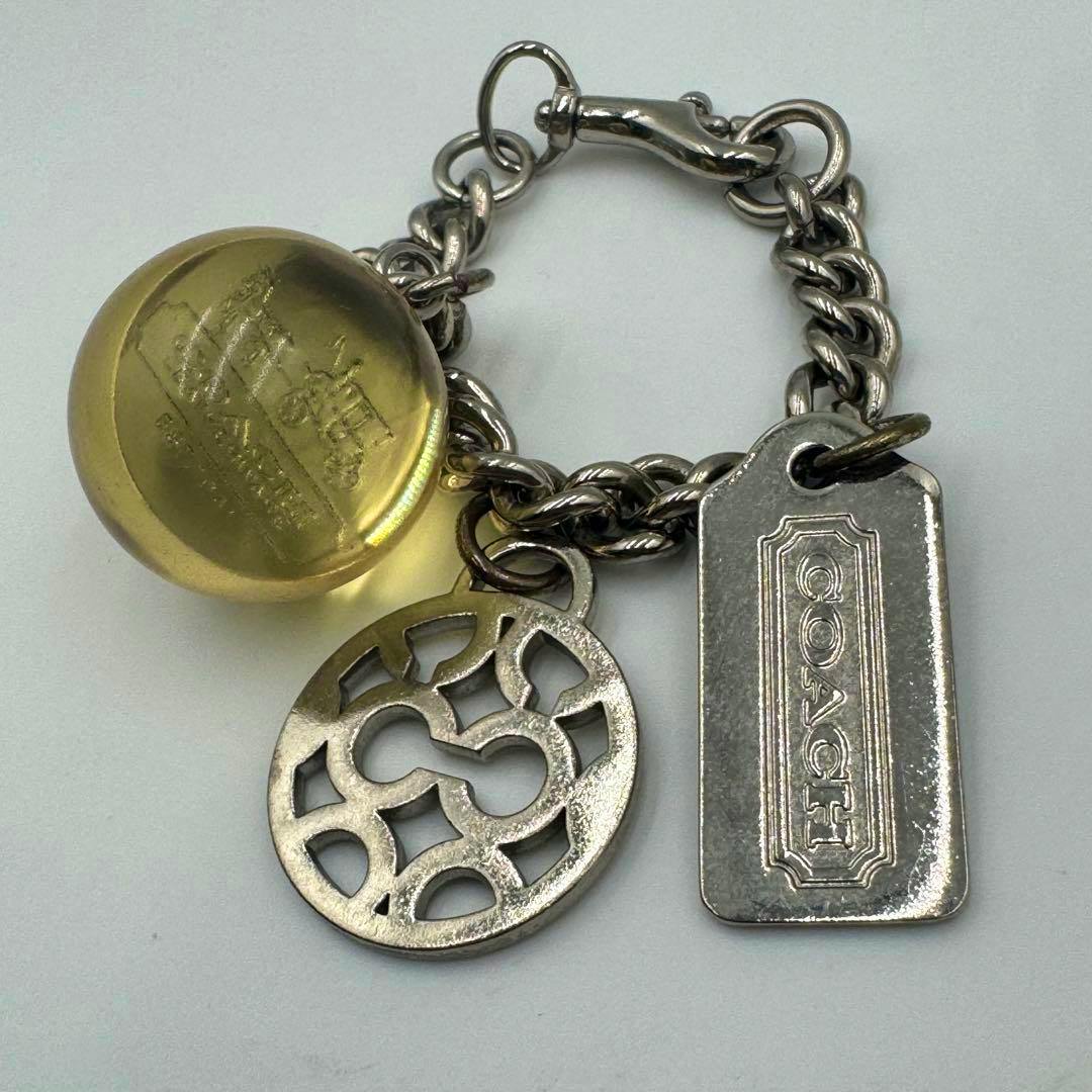 A Coach bag charm with a silver-toned chain and clasp, a rectangular Coach tag, a round openwork medallion, and a yellow domed charm.