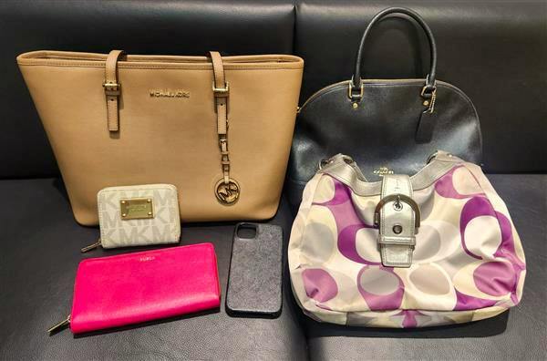 Six pre-owned branded bags and small accessories arranged on a black surface, including a tan tote, a black dome bag, a purple-patterned shoulder bag, a pink zipper wallet, a small white wallet, and a phone case.
