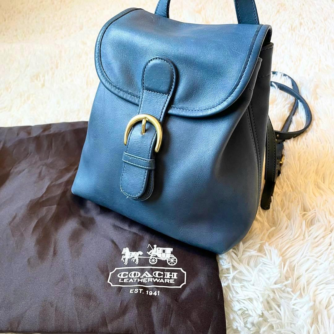 A blue Coach mini backpack (cow leather) shown on a light fur surface with a brown Coach dust bag beneath it.