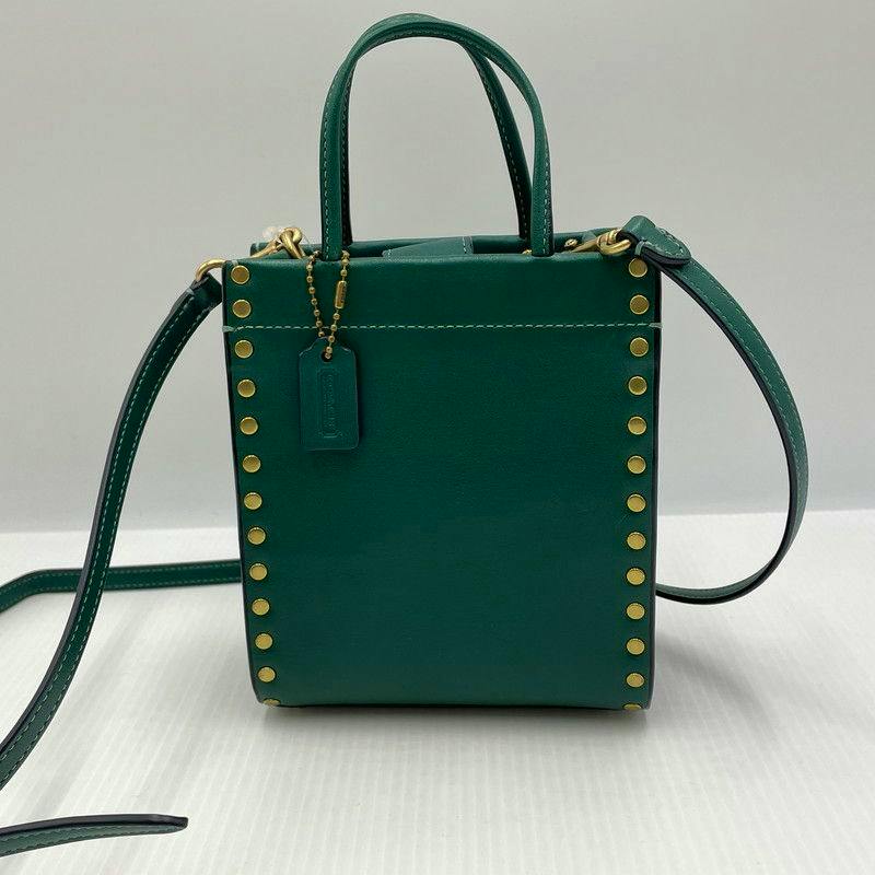 Front view of a green COACH Mini Casing Tote bag with gold rivets, two top handles, a long shoulder strap, and a hanging tag.