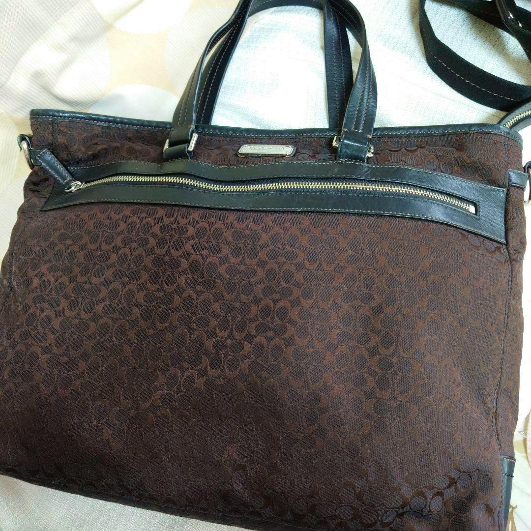 Front view of a brown Coach mini signature canvas business bag with leather trim, top handles, and a front zip pocket.