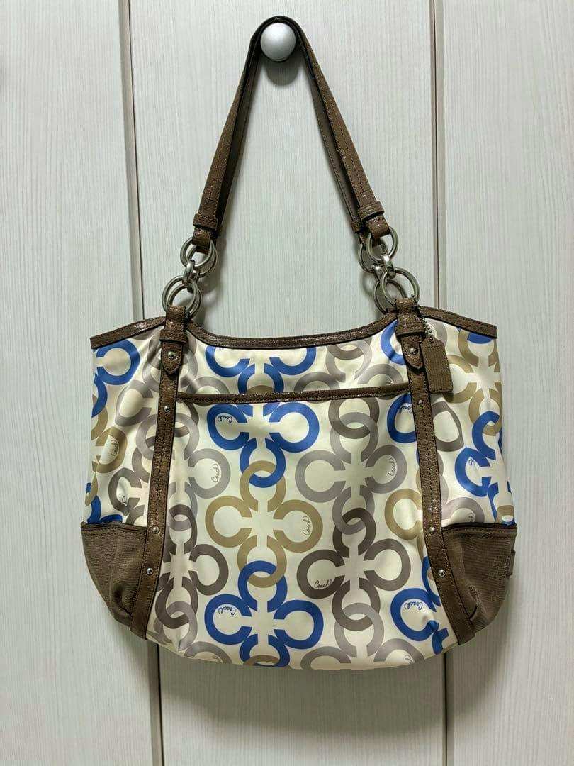 Multicolor COACH canvas shoulder bag with blue, tan, and gray interlocking C pattern and brown straps hanging on a hook against a light background.
