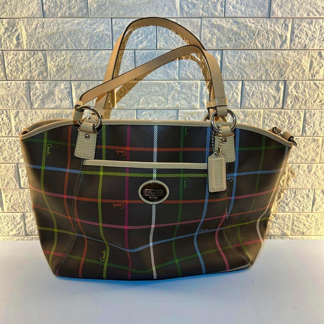 Front view of a brown COACH canvas tote bag with a multicolor check pattern and beige handles against a tiled wall.