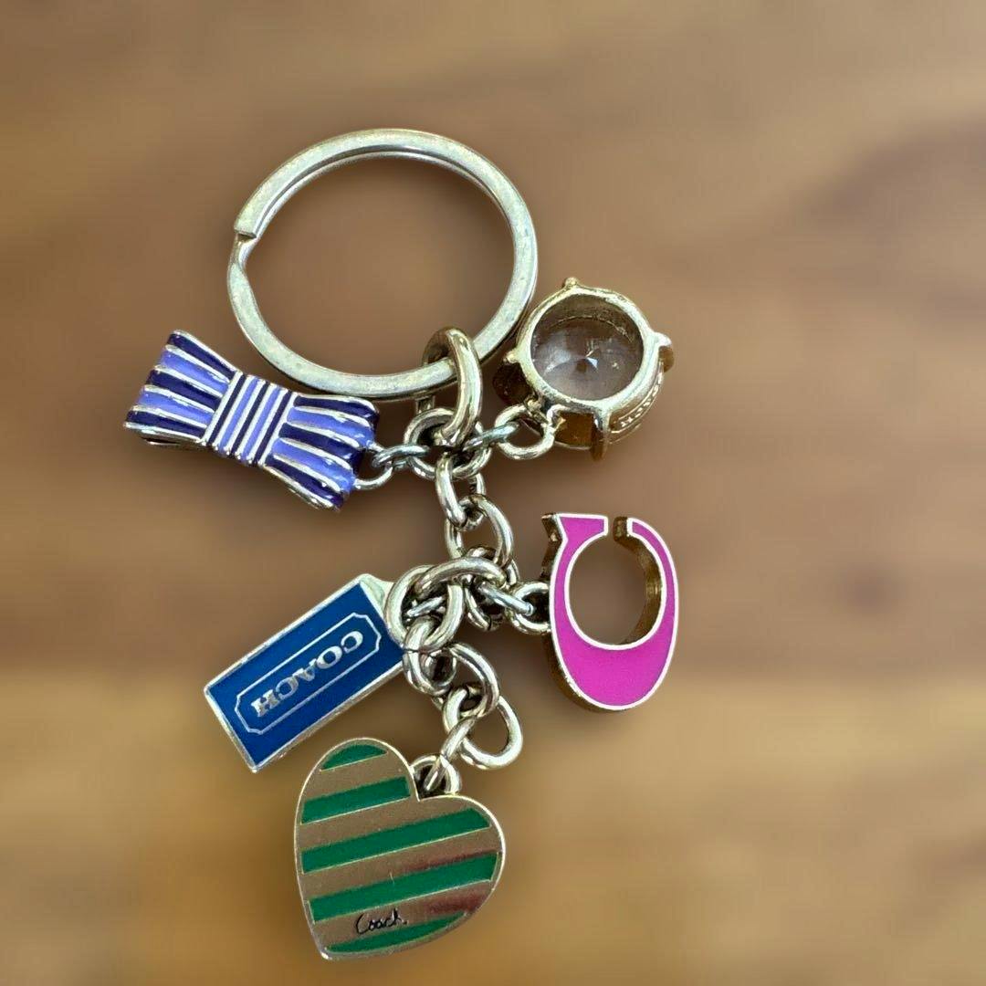 Multicolor COACH keychain with enamel charms — striped heart, blue COACH tag, pink C-shaped charm, purple bow, and a clear faceted stone on a metal key ring.