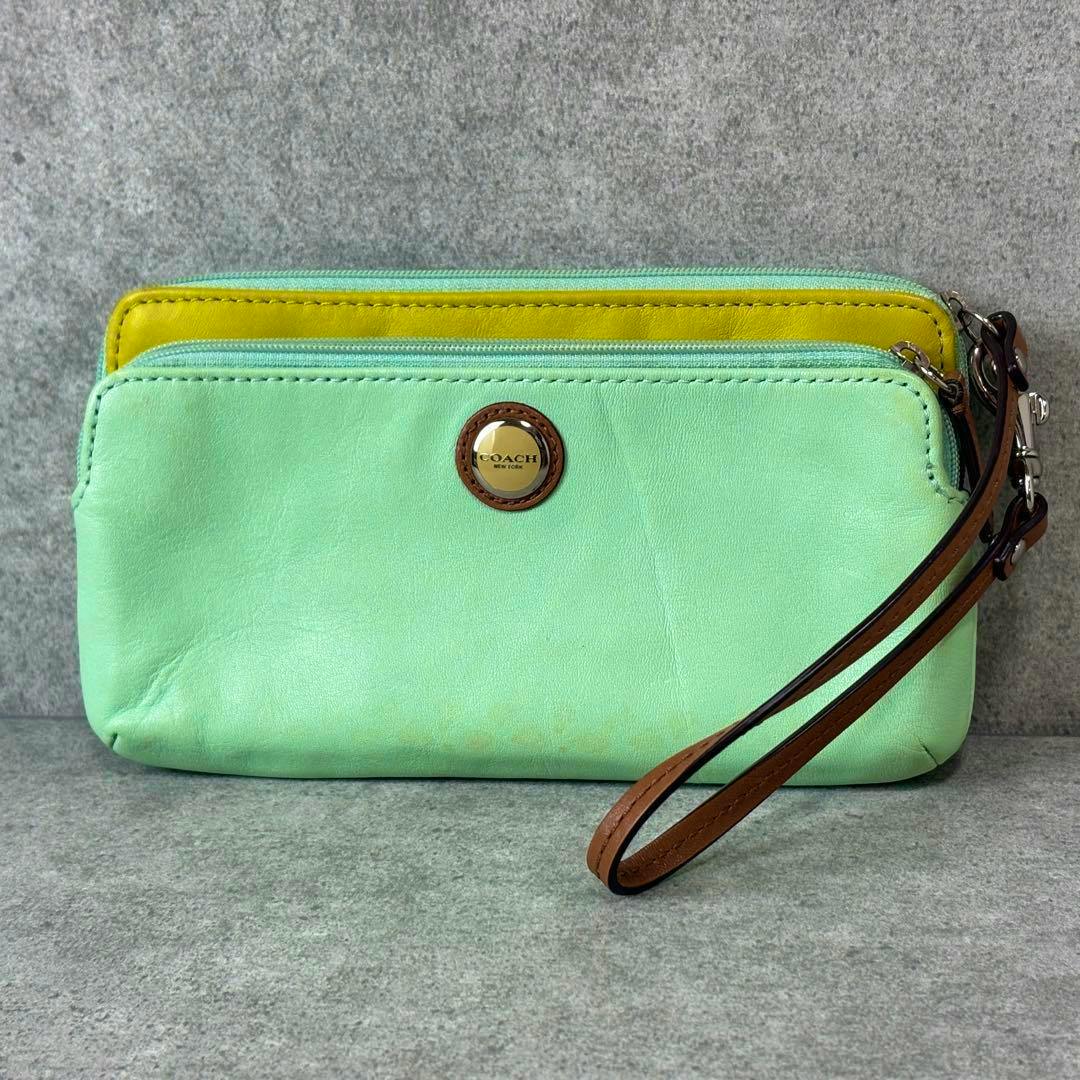 Front view of a lime-green and yellow Coach leather wristlet pouch with a brown wrist strap and round Coach logo emblem.