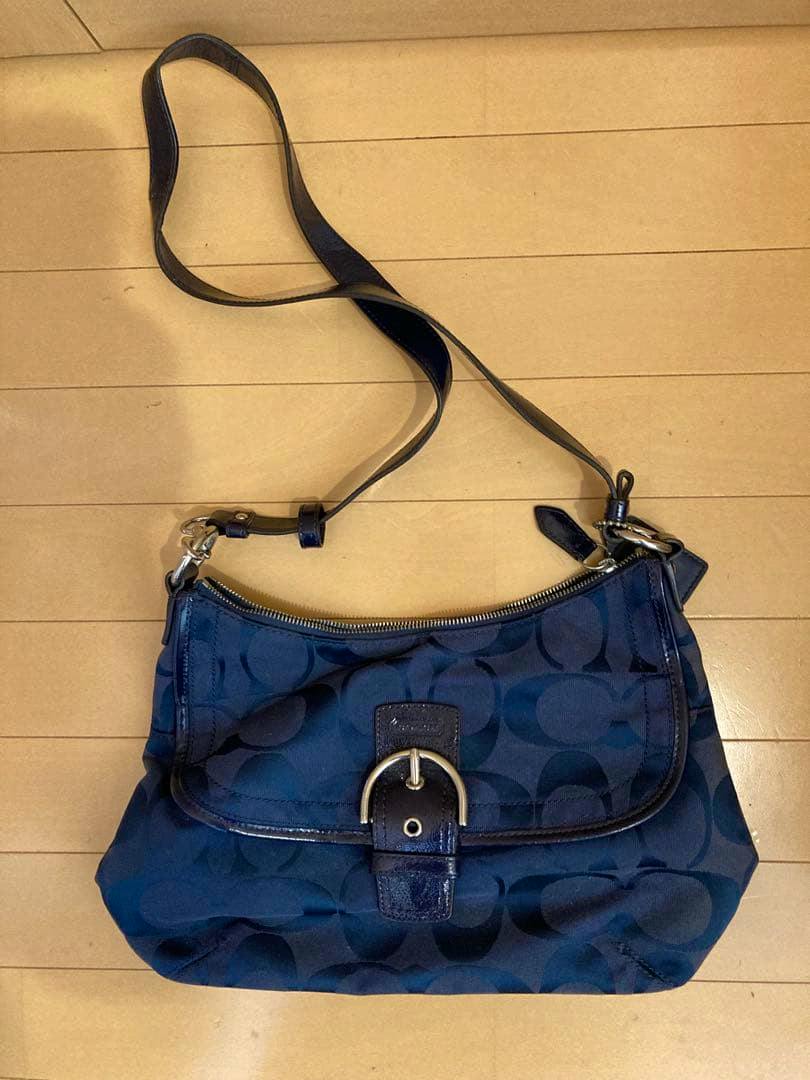 Front view of a navy Coach nylon shoulder bag with dot pattern, flap with metal buckle and adjustable leather strap on a wooden floor.