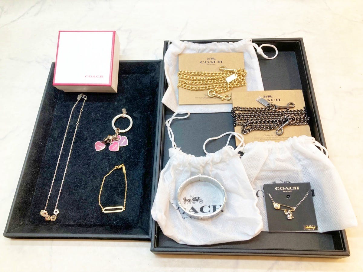 An assortment of Coach necklaces, chains, bracelets, and other accessories arranged on trays and in branded pouches.