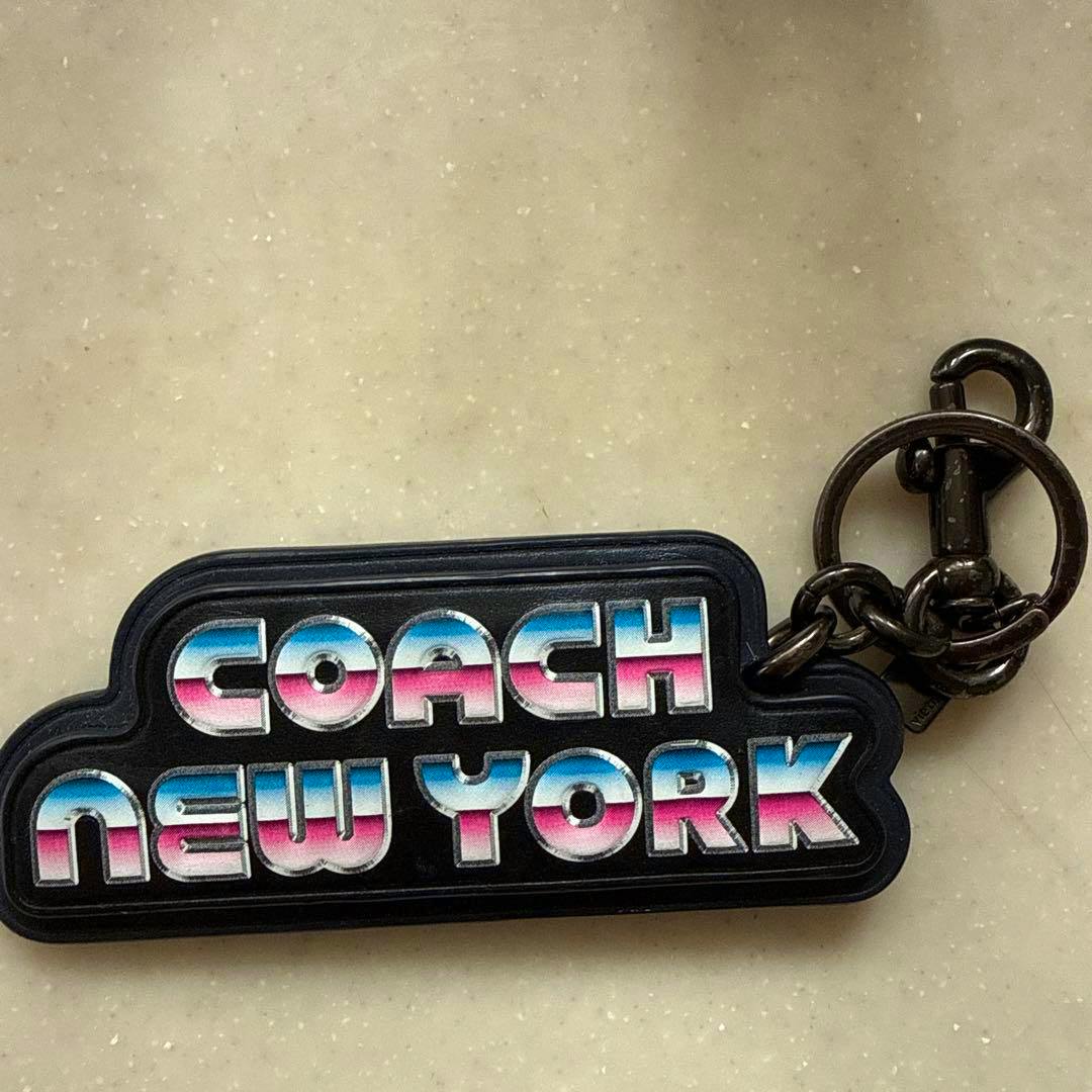 Front view of a COACH NEW YORK keychain keyring showing multicolored "COACH NEW YORK" text on a black leather tag with metal rings.