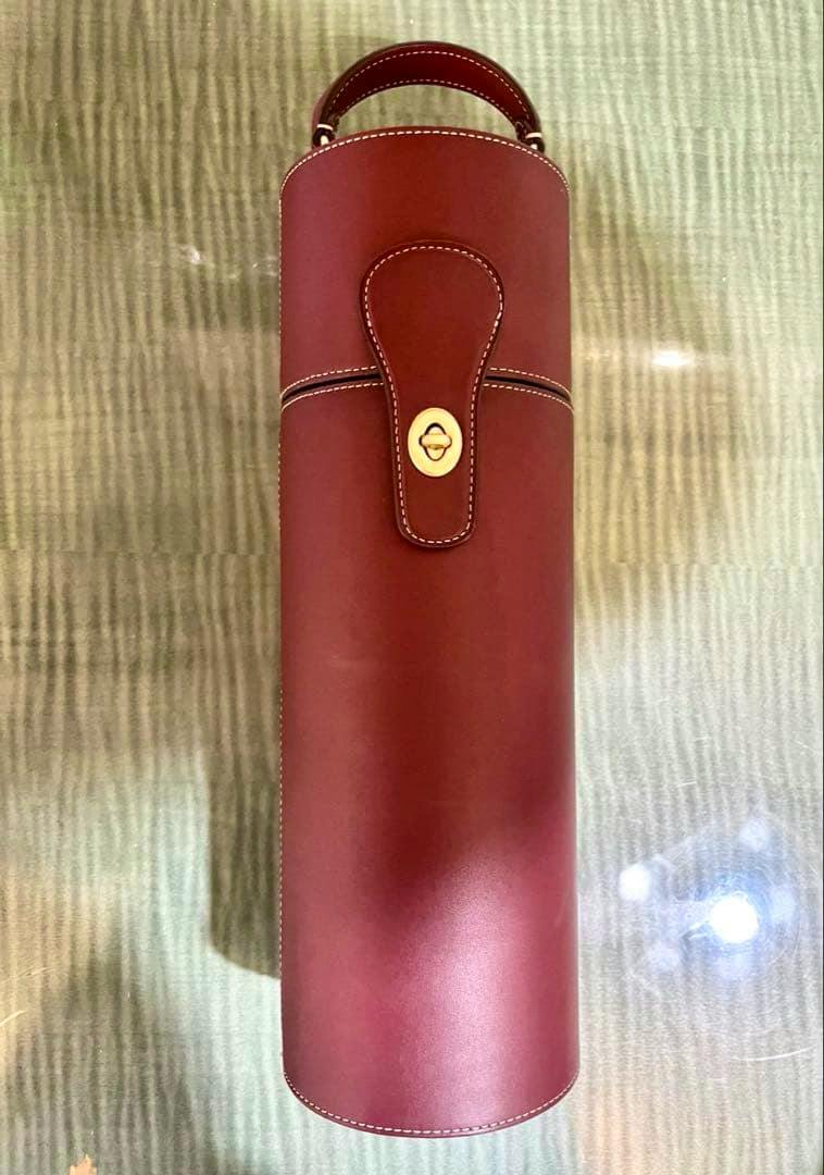 Red cylindrical leather wine bottle case with top handle and front strap with gold turn-lock shown on a textured surface.