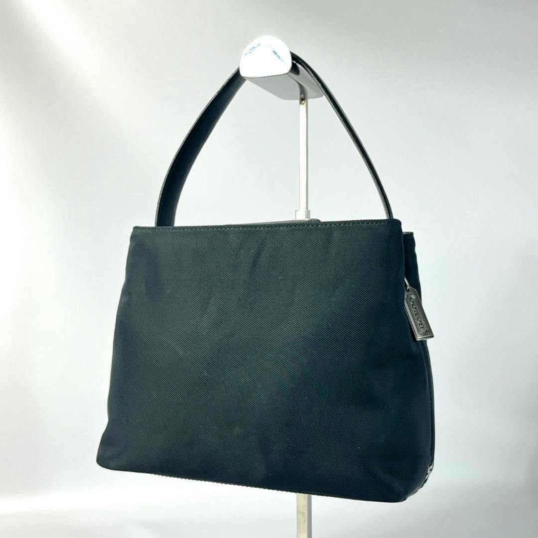 Black Coach nylon handbag with a leather handle and metal charm displayed on a stand.