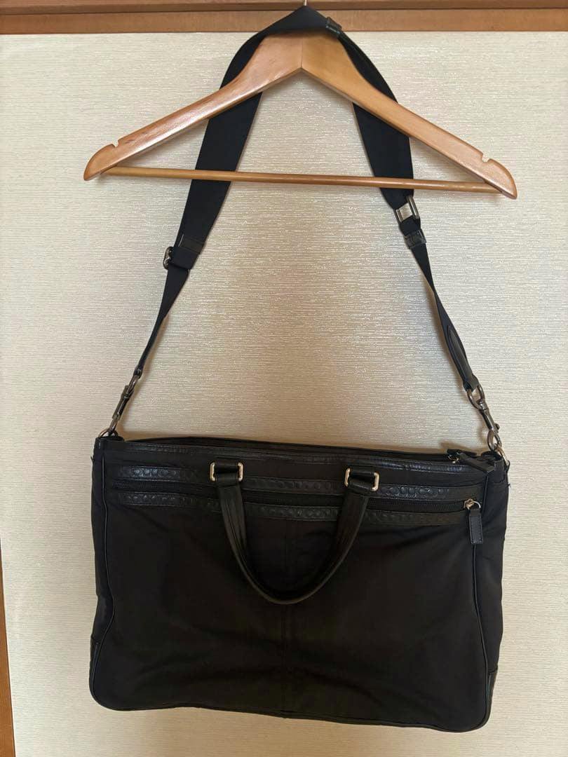 Black Coach nylon and leather business bag hanging from a wooden hanger, showing the front, handles, and adjustable shoulder strap.