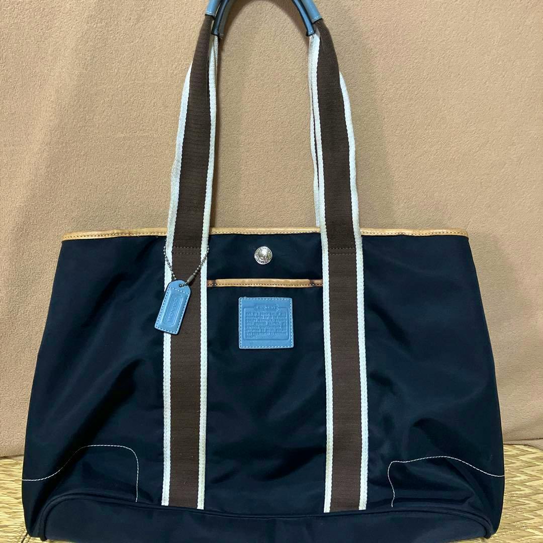 Front view of the COACH black nylon tote bag with brown-and-white striped handles, blue leather logo patch and tag, and tan top trim.