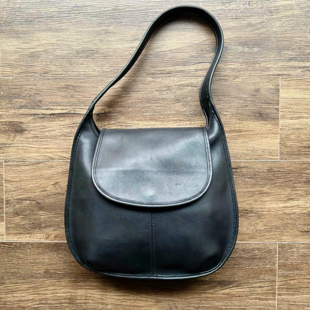 Front view of a black leather Coach Ergo Flap shoulder bag with its curved flap visible on a wood floor.