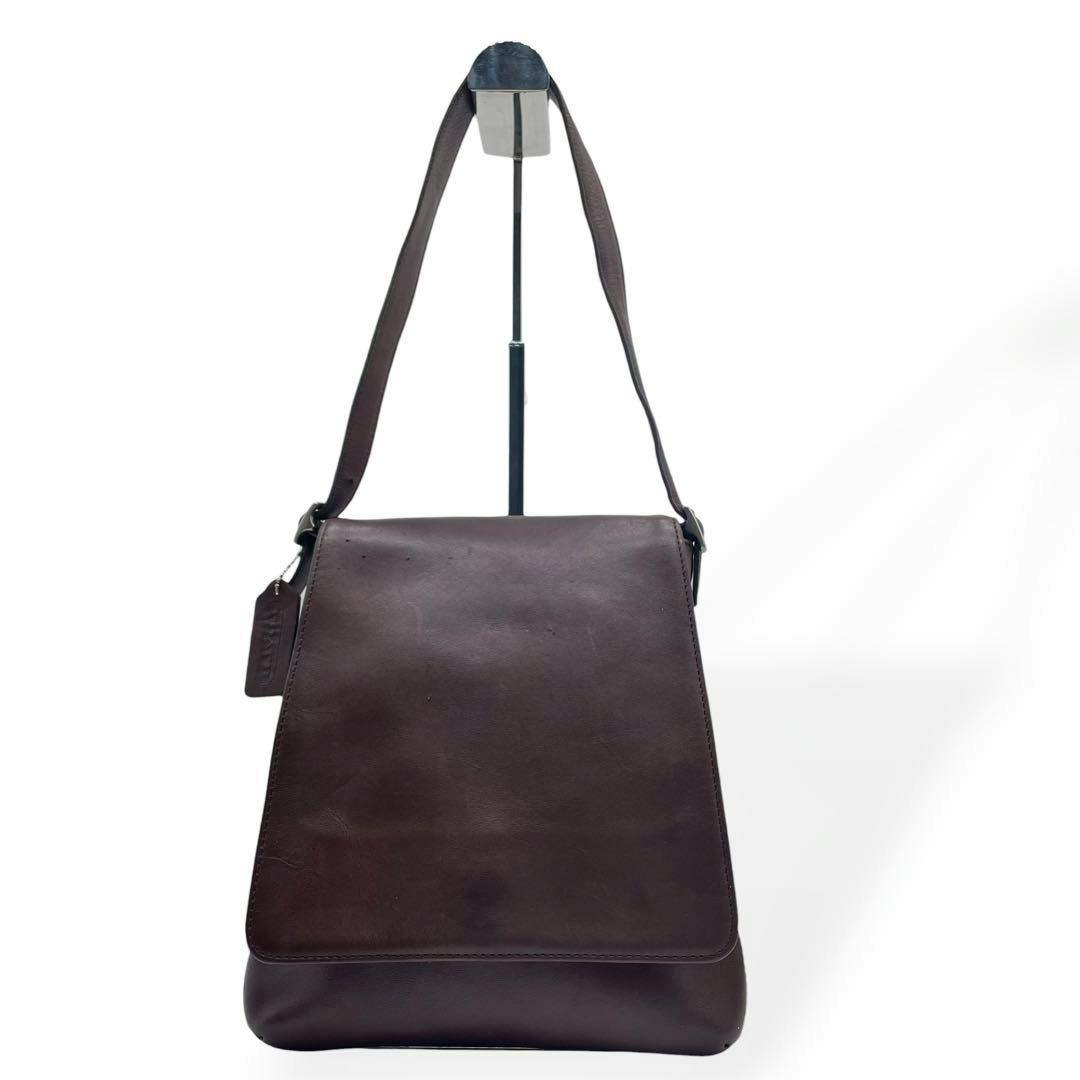 Front view of a brown Coach shoulder bag with flap hanging on a stand against a white background.