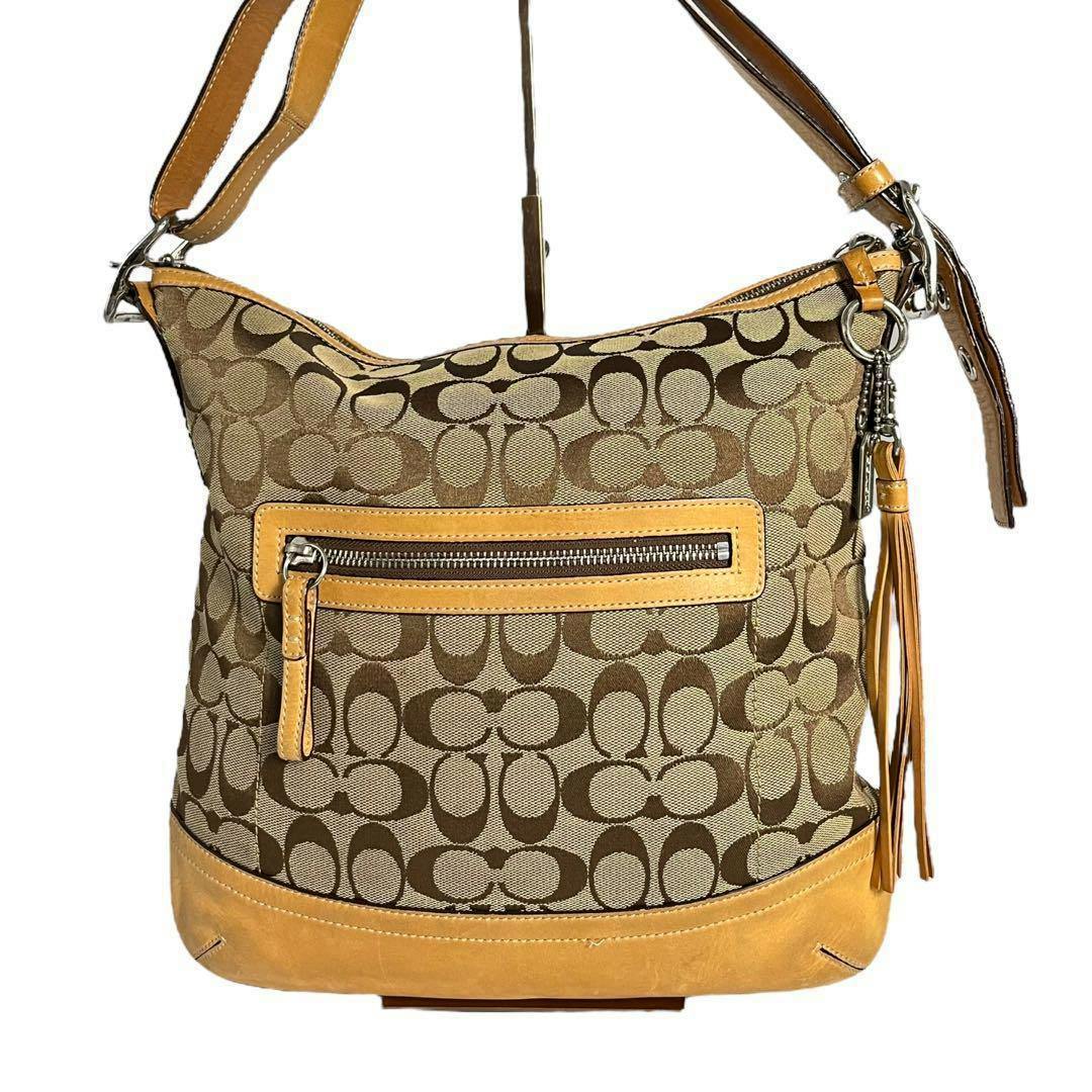 Front view of a COACH canvas shoulder bag with brown monogram pattern, tan leather trim, front zip pocket, shoulder strap, and leather tassel.