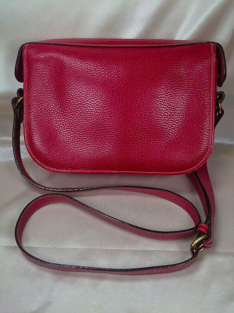 Front view of a red COACH cow leather shoulder bag with long strap and brass hardware on a light fabric background.