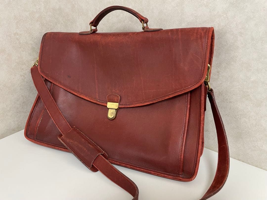 Brown leather briefcase with top handle, front flap closure, and adjustable shoulder strap on a light surface.
