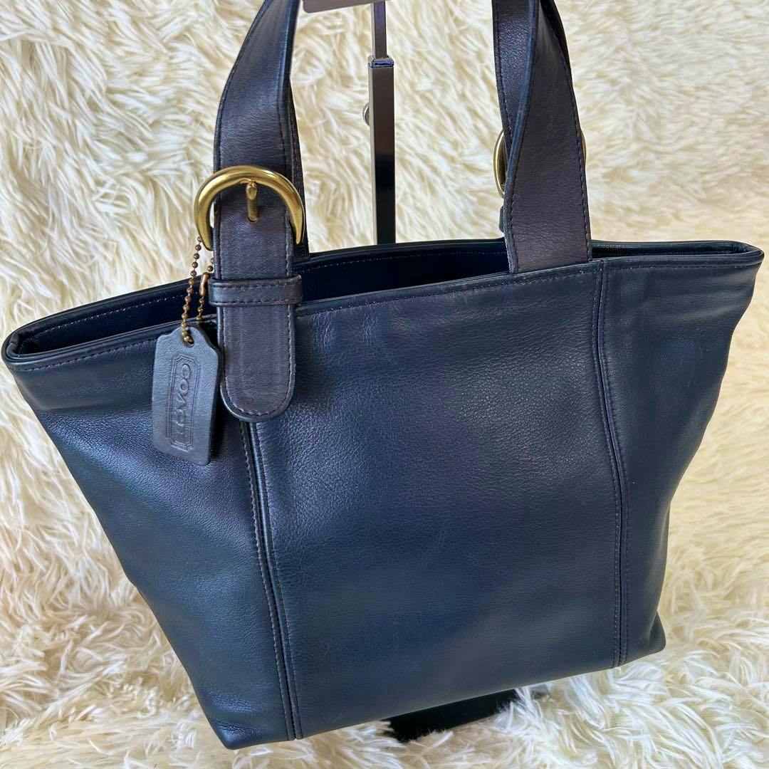 Black leather Coach handbag with two top handles and a hanging leather tag shown against a cream fur background.