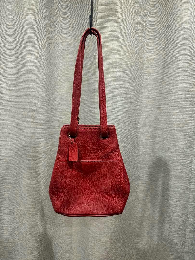 Red COACH leather handbag hanging by its straps against a neutral curtain background.