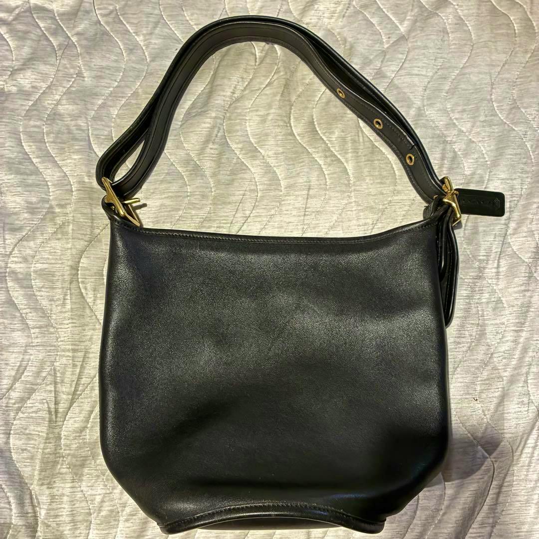 Black cow leather COACH shoulder bag laid flat showing the front, adjustable shoulder strap, and leather hang tag.