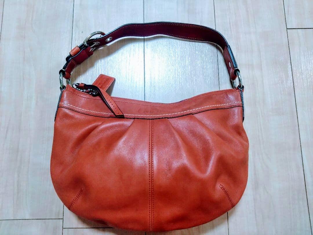 Orange leather shoulder bag laid flat on a wooden floor, front view with the shoulder strap attached.