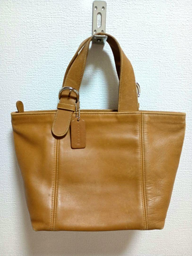 Front view of the camel COACH cow leather tote bag hanging on a wall, showing handles and a leather tag.