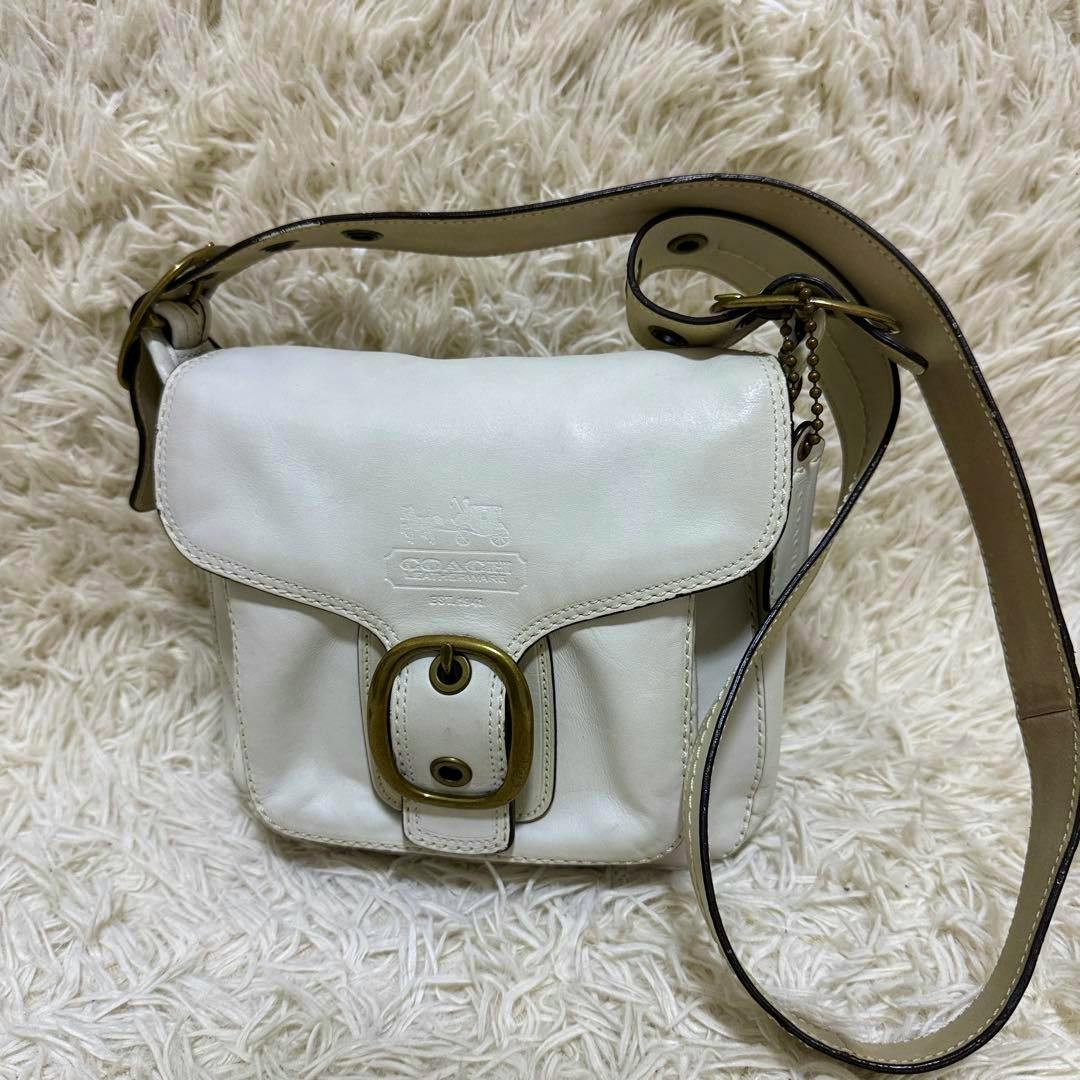 White COACH cow leather shoulder bag with front buckle and adjustable strap resting on a soft rug.