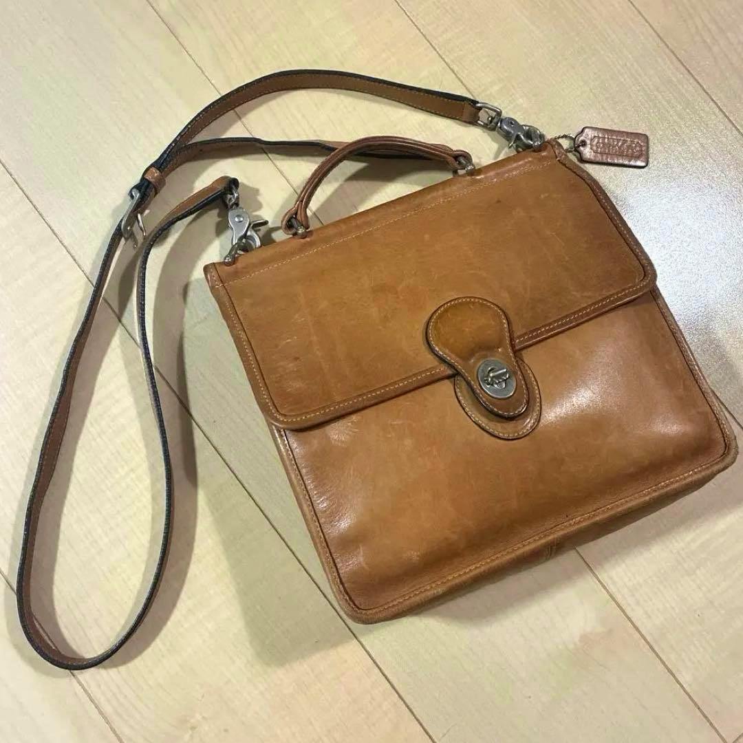 Brown cow leather Coach 2-way shoulder bag with flap and clasp, top handle and long strap lying on a light wood floor.