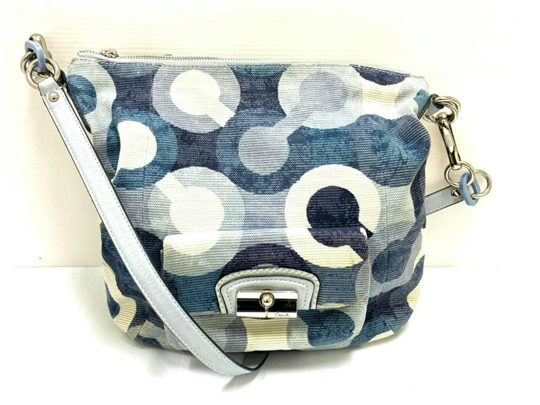 Front view of a Coach canvas shoulder bag with blue-and-white circular op-art pattern and a light-colored leather strap.