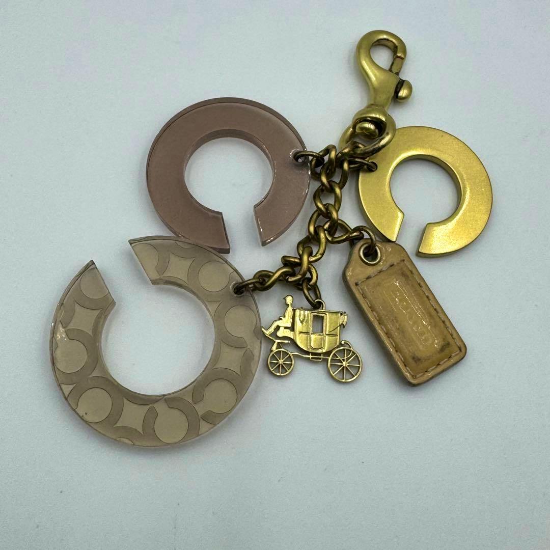 Coach bag charm (zinc alloy) showing a gold-toned carriage charm, leather tag, chain, clasp, and circular op-art charms on a white background.