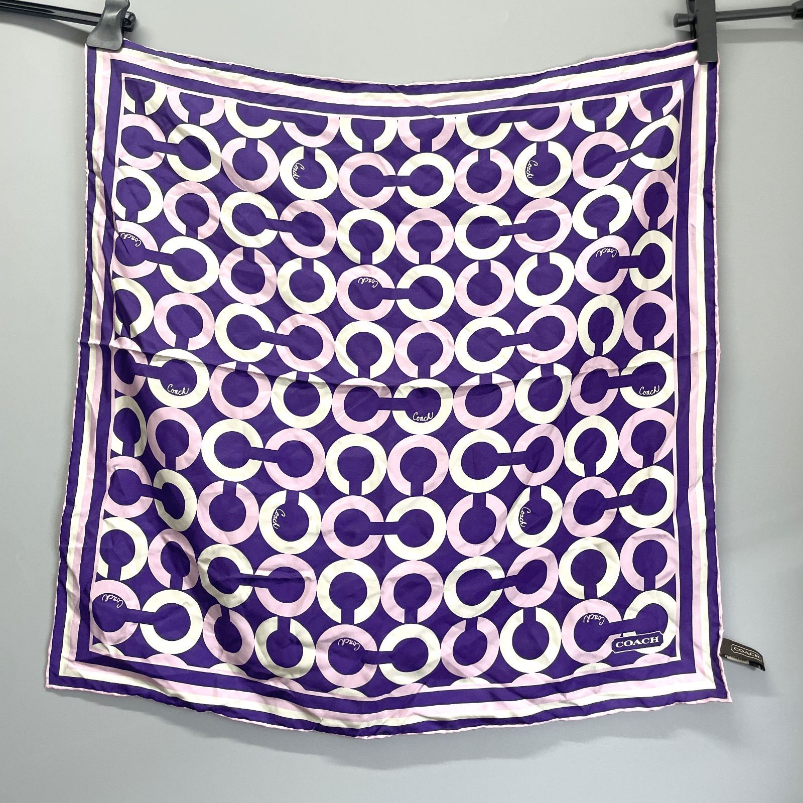 Square 65×65 cm Coach op art silk scarf in purple and white with interlocking circular pattern, clipped and hanging against a gray background.